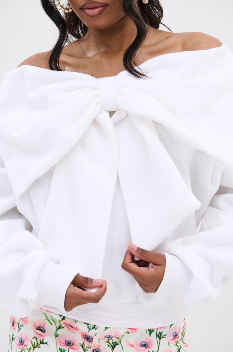 A woman wears THE BEST BOW SWEATER, a white off-the-shoulder top with a large bow applique at the chest, paired with floral print pants and holding the sides of her top.