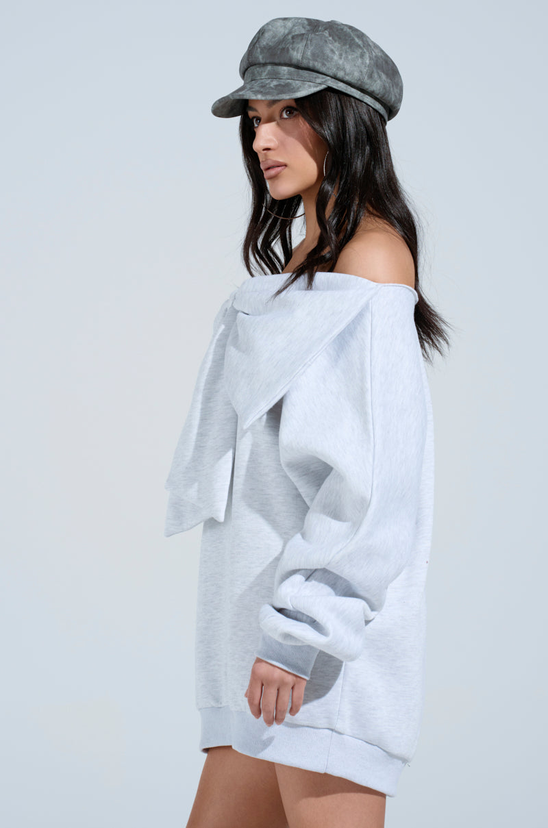 A woman with long dark hair stands sideways, gazing right against a plain background, wearing an off-the-shoulder "WHO ME? BOW FRONT SWEATSHIRT DRESS" in grey with a large bow front and a matching cap.