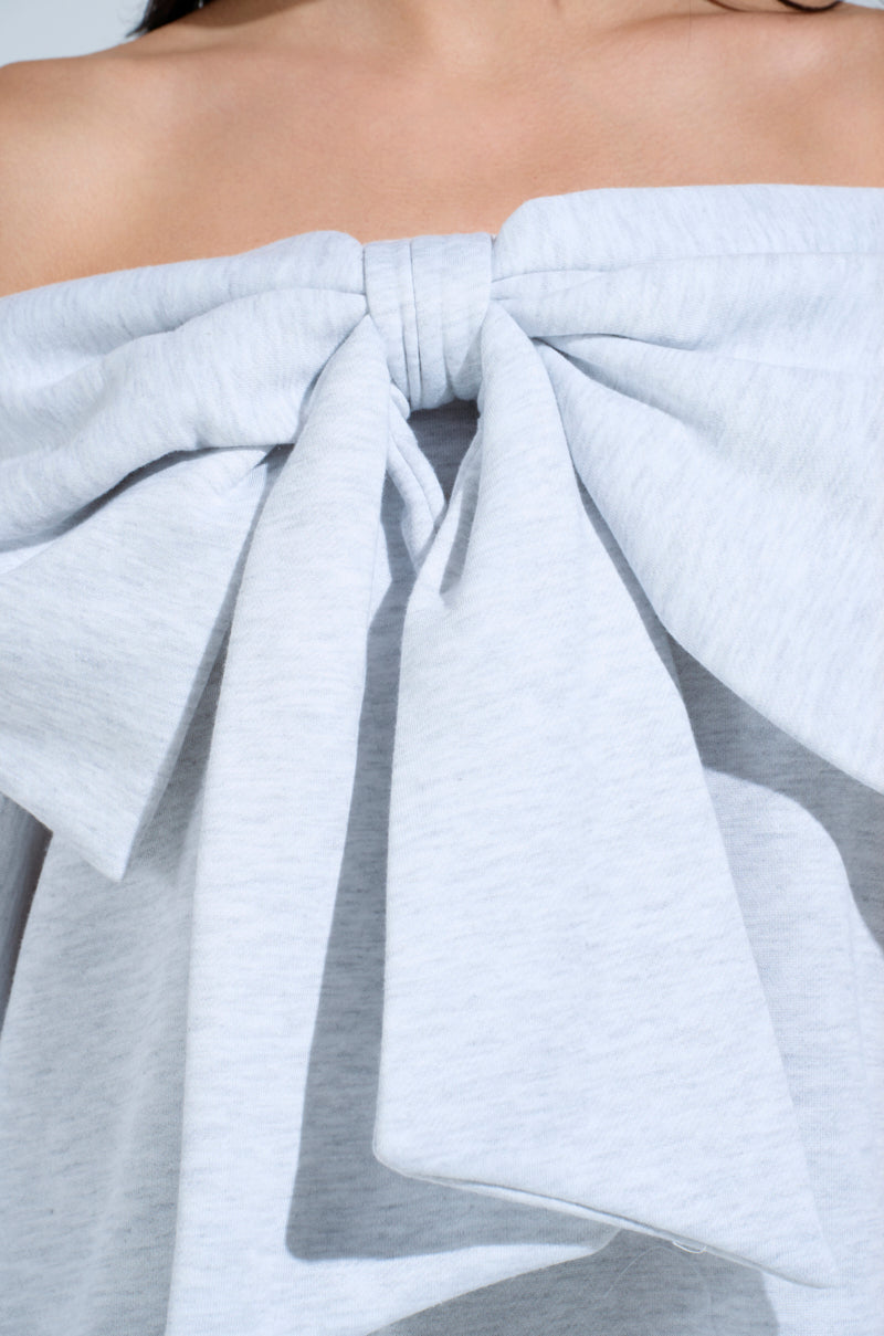 Close-up of the WHO ME? Bow Front Sweatshirt Dress in light gray, featuring an elegant off-shoulder design with a large, elaborate bow at the chest. The garment has a soft textured fabric, with only part of the neck and collarbones visible above it.