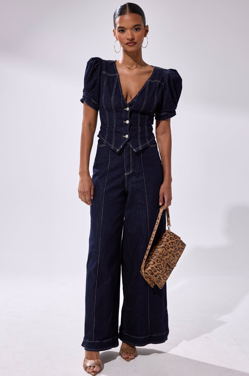 A woman wears the CHIC AF DENIM JUMPSUIT featuring a fitted short-sleeve button-up top and wide-leg pants. She completes her look with hoop earrings, heeled sandals, and a leopard print handbag.
