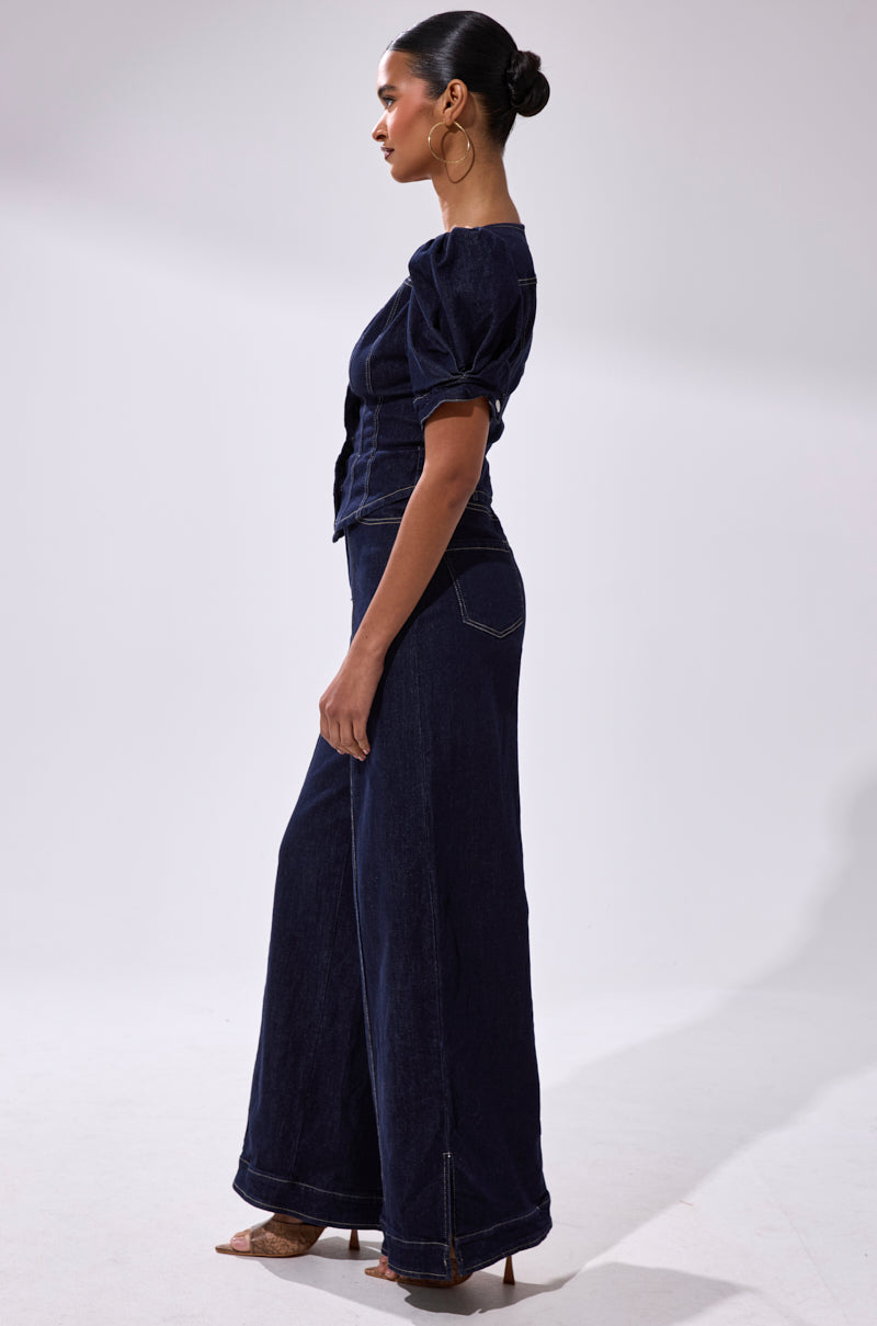 A woman stands in profile wearing the CHIC AF DENIM JUMPSUIT, a dark indigo one-piece featuring puffed short sleeves and wide-leg pants. She pairs it with hoop earrings, heels, and a sleek bun against a light background.