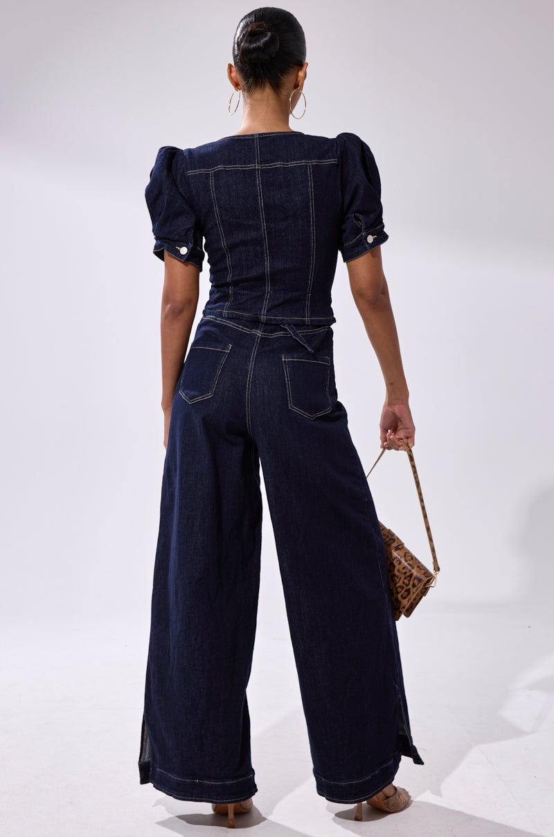 A woman faces away, wearing the CHIC AF DENIM JUMPSUIT in dark indigo with puffed short sleeves and wide-leg pants. She holds a small leopard-print handbag and has her hair styled in a sleek bun.