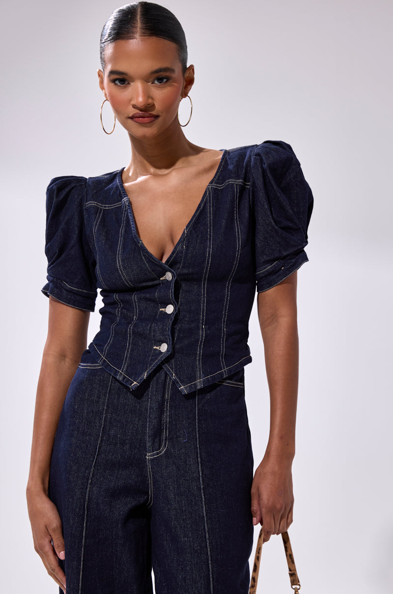 A woman stands against a plain light background, wearing the CHIC AF DENIM JUMPSUIT in dark indigo with puffed short sleeves, a deep V-neck, and front buttons. She accessorizes with large hoop earrings and holds a small handbag.