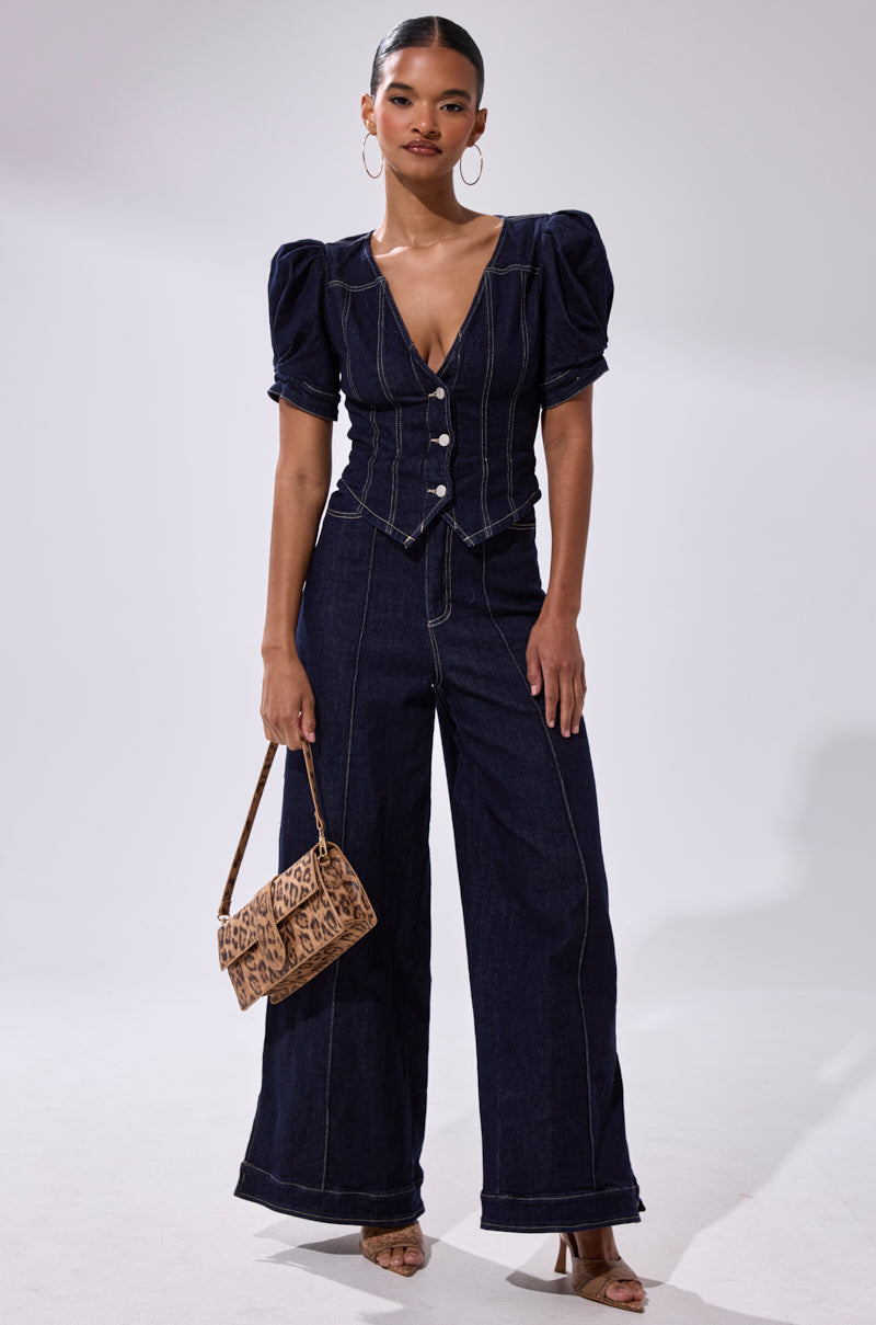 A woman models the CHIC AF DENIM JUMPSUIT—dark indigo with puffed short sleeves, a deep V-neck, and wide-leg pants—styled with hoop earrings, heeled sandals, and a leopard print handbag.