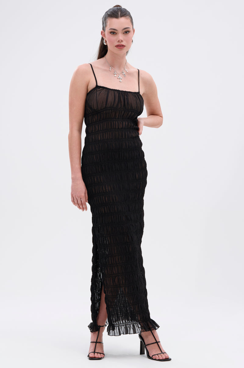 A woman poses against a white background in the ON A BEACH OR A NIGHT OUT MESH MAXI DRESS—a sheer, ruched black mesh, ankle-length maxi with thin straps. She styles it with strappy black heels, a silver necklace, and a sleek updo for day-to-night appeal.