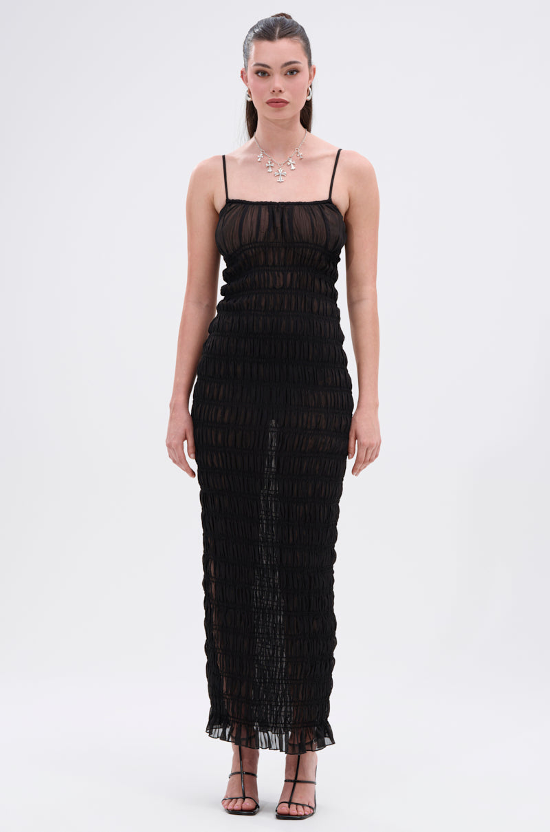 A woman faces forward in the ON A BEACH OR A NIGHT OUT MESH MAXI DRESS, a figure-hugging black mesh maxi with thin straps. She pairs it with black sandals, a silver necklace and earrings. Her straight brown hair is pulled back, against a plain white background.