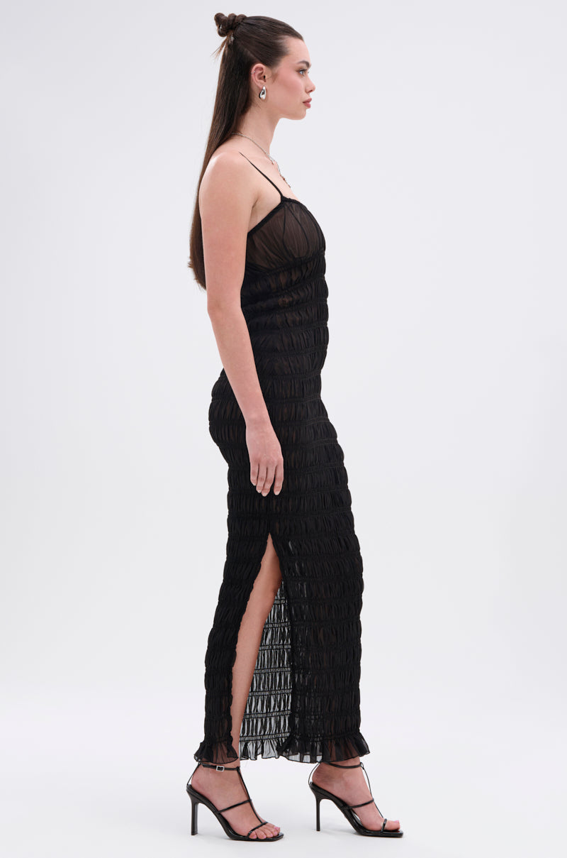 A woman in profile wears the ON A BEACH OR A NIGHT OUT MESH MAXI DRESS, featuring textured ruching and a side slit, paired with strappy black heels. Her long hair is styled half-up against a plain background—an easy day-to-night look.