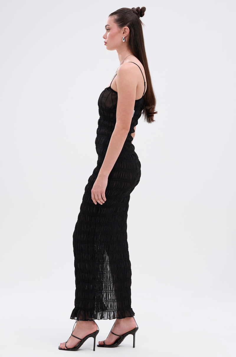 A woman stands in profile against a white background, wearing the ON A BEACH OR A NIGHT OUT MESH MAXI DRESS—a sleek black sleeveless piece with thin straps and textured fabric—paired with black strappy high-heeled sandals.