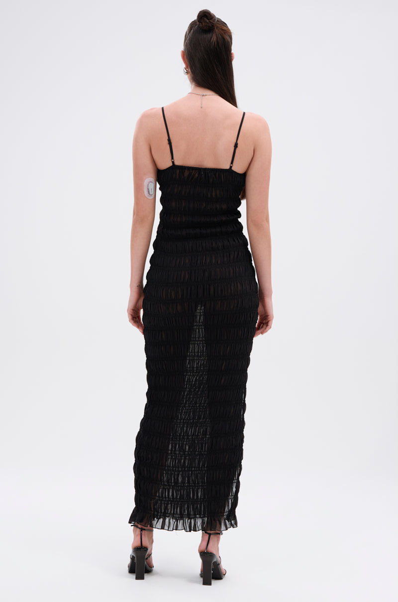 A woman with long brown hair wears the ON A BEACH OR A NIGHT OUT MESH MAXI DRESS and heels, standing with her back to the camera. A medical patch is visible on her upper left arm, showing this dress as a versatile day-to-night option.