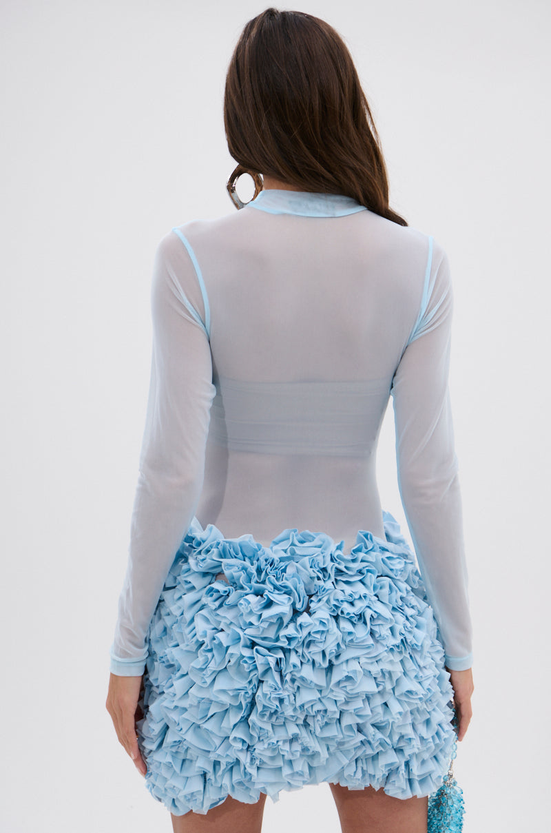 A woman with long brown hair, seen from the back, wears the KELYN MESH MINI DRESS IN BLUE—a sheer fitted top and layered blue skirt. She holds a small blue beaded bag in her left hand, completing her elegant look.