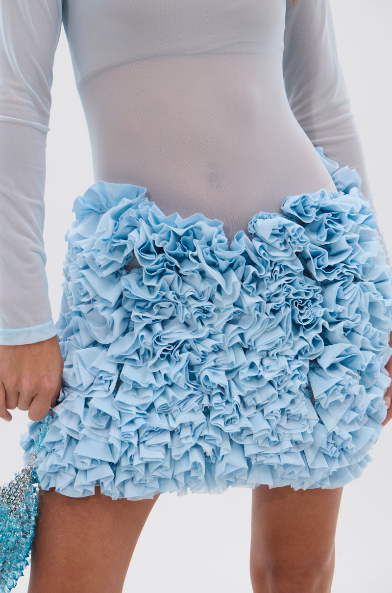 Wearing the KELYN MESH MINI DRESS IN BLUE, featuring light mesh fabric with ruffled layers and a short skirt, a person holds a beaded blue handbag. The photo frames only their torso and upper legs.