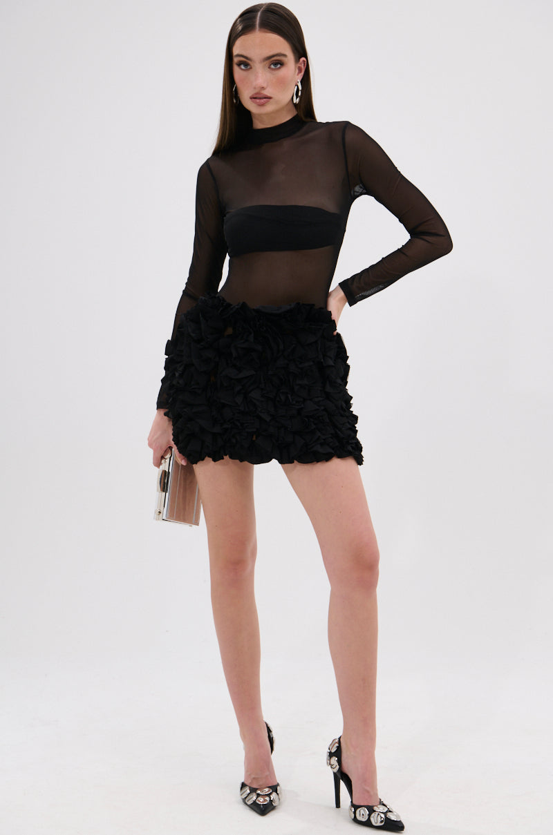 A woman radiates elegance in the KELYN MESH MINI DRESS IN BLACK, featuring sheer long sleeves and ruffle details. She accessorizes with a beige clutch and black heels with silver accents, set against a white background.