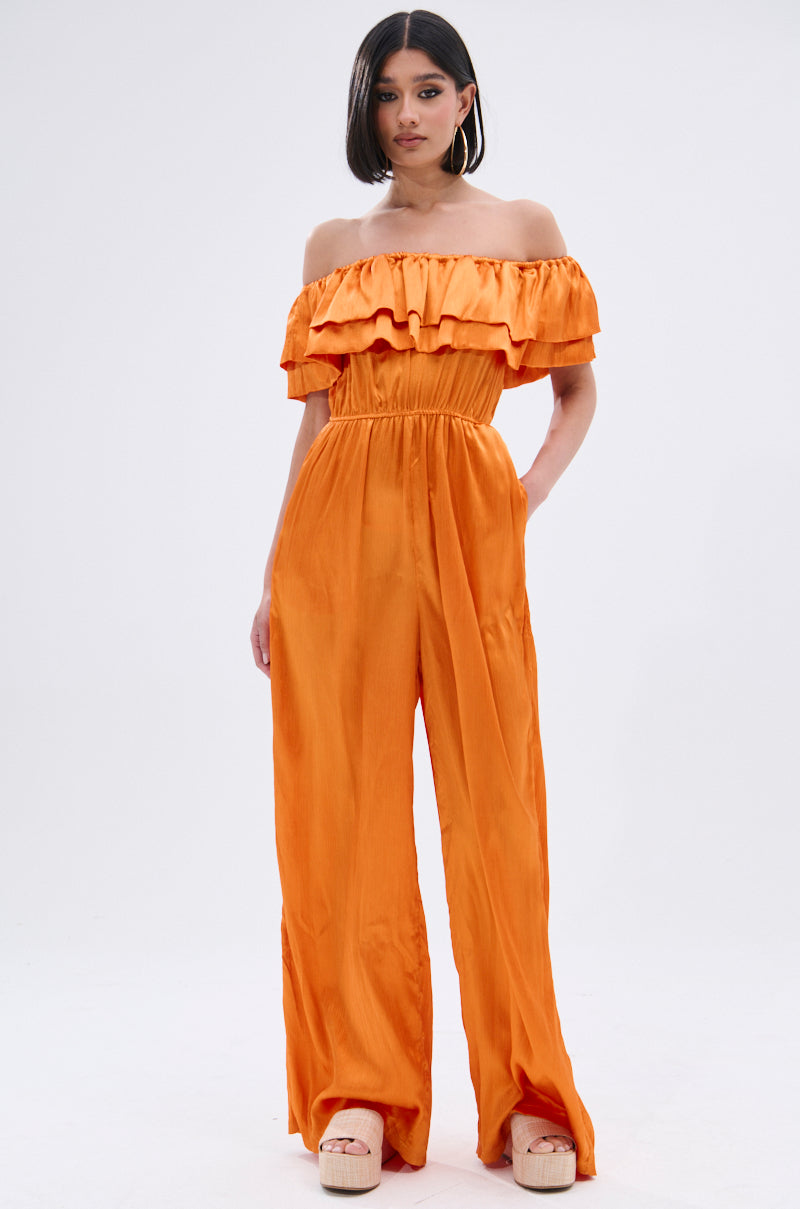 A woman poses against a light background in the BYE BYE WINTER BLUES OFF THE SHOULDER SATIN JUMPSUIT, featuring vibrant orange satin, ruffle details, wide-leg pants, and platform sandals. She wears large hoop earrings with straight shoulder-length dark hair.
