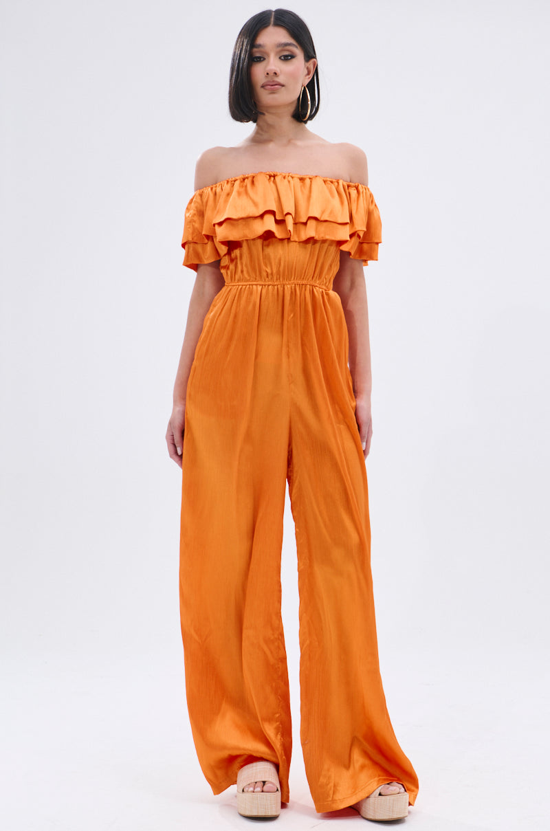 A woman with straight dark hair poses against a white background in the BYE BYE WINTER BLUES OFF THE SHOULDER SATIN JUMPSUIT, featuring ruffle detailing and wide-leg pants, styled with platform sandals.