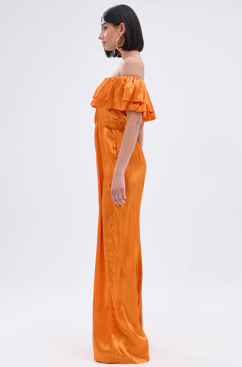 A woman poses in profile against a plain light background, wearing the BYE BYE WINTER BLUES OFF THE SHOULDER SATIN JUMPSUIT with ruffle details and wide-leg pants. She has chin-length straight hair and hoop earrings.