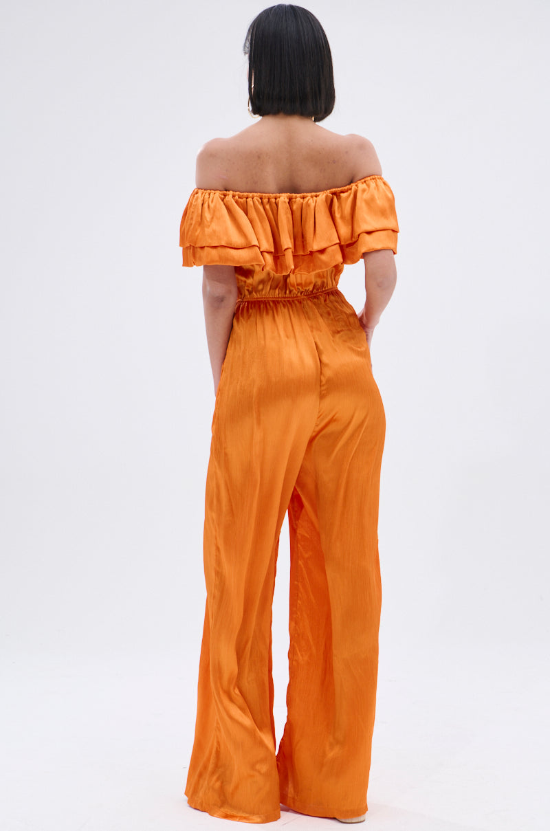 A woman with a bob haircut faces away, modeling the BYE BYE WINTER BLUES OFF THE SHOULDER SATIN JUMPSUIT in vibrant orange with ruffle details and wide-leg pants against a plain white background.