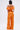 A woman with a bob haircut faces away, modeling the BYE BYE WINTER BLUES OFF THE SHOULDER SATIN JUMPSUIT in vibrant orange with ruffle details and wide-leg pants against a plain white background.