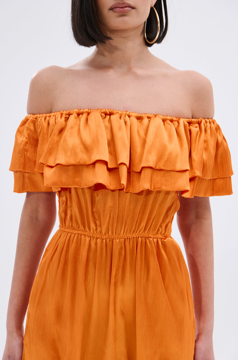 A woman models the BYE BYE WINTER BLUES OFF THE SHOULDER SATIN JUMPSUIT in bright orange, featuring ruffle details and a gathered waist. Her straight chin-length hair and large hoop earrings complete the look. Cropped from shoulders to knees.