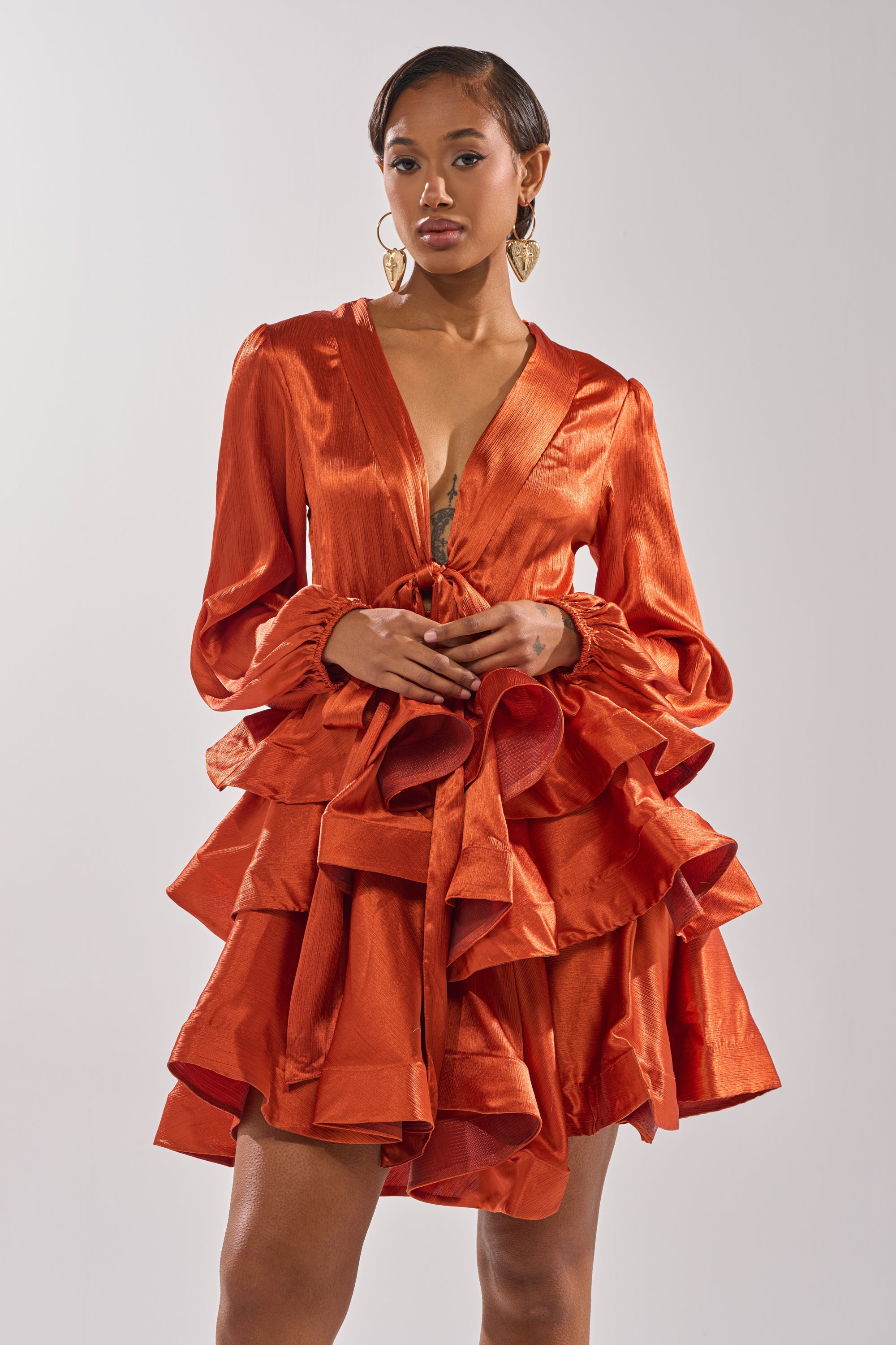 A woman in the ARIANA SATIN RUFFLED MINI DRESS, with long sleeves, a deep V-neckline, and layered ruffles in shiny burnt orange, stands confidently against a plain background.