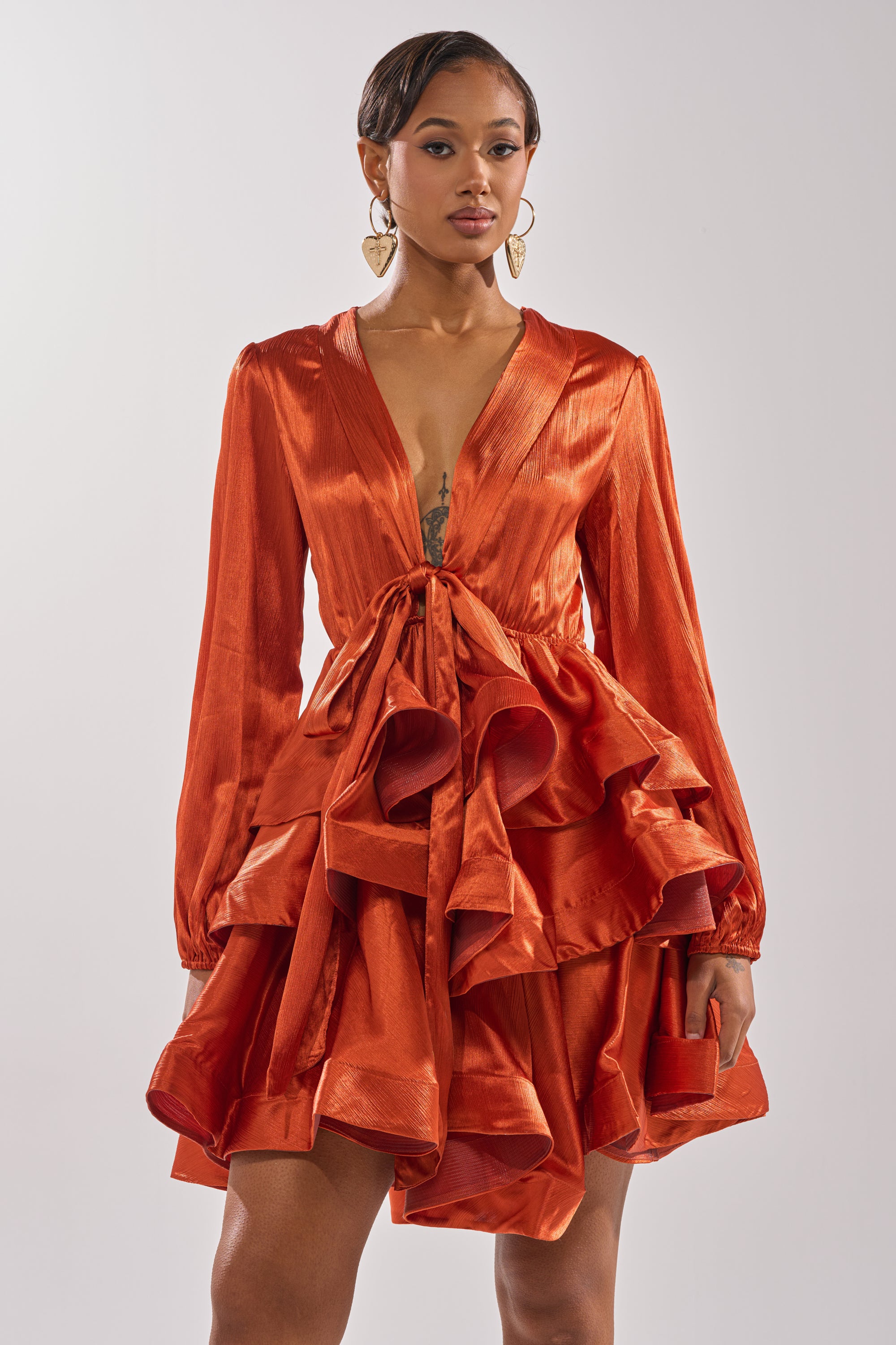 A woman poses against a plain background in the ARIANA SATIN RUFFLED MINI DRESS—a shiny burnt orange mini dress with long sleeves, deep V-neck, dramatic skirt ruffles, and a matching purse—ideal for chic evening events.