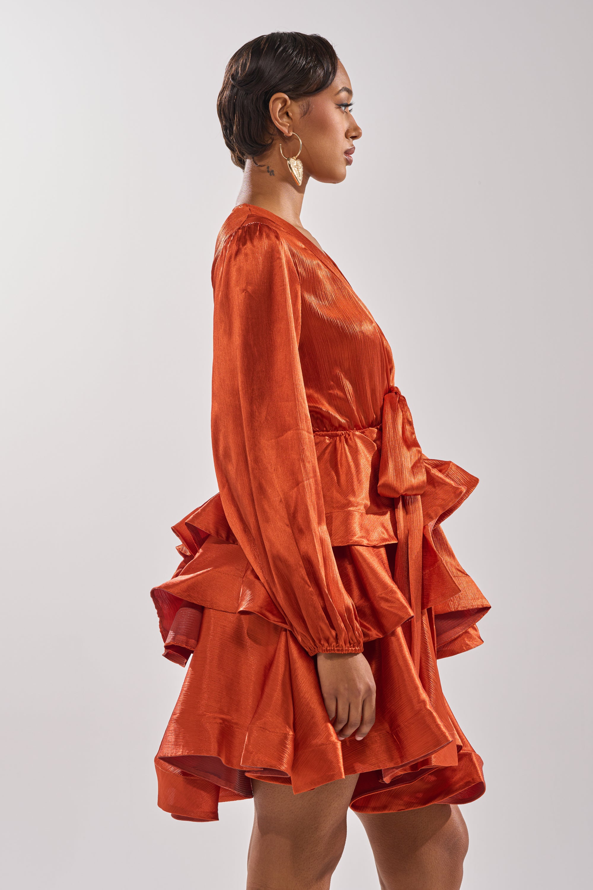 A woman with short hair and large hoop earrings poses in profile against a plain light background, wearing the ARIANA SATIN RUFFLED MINI DRESS with a waist tie.