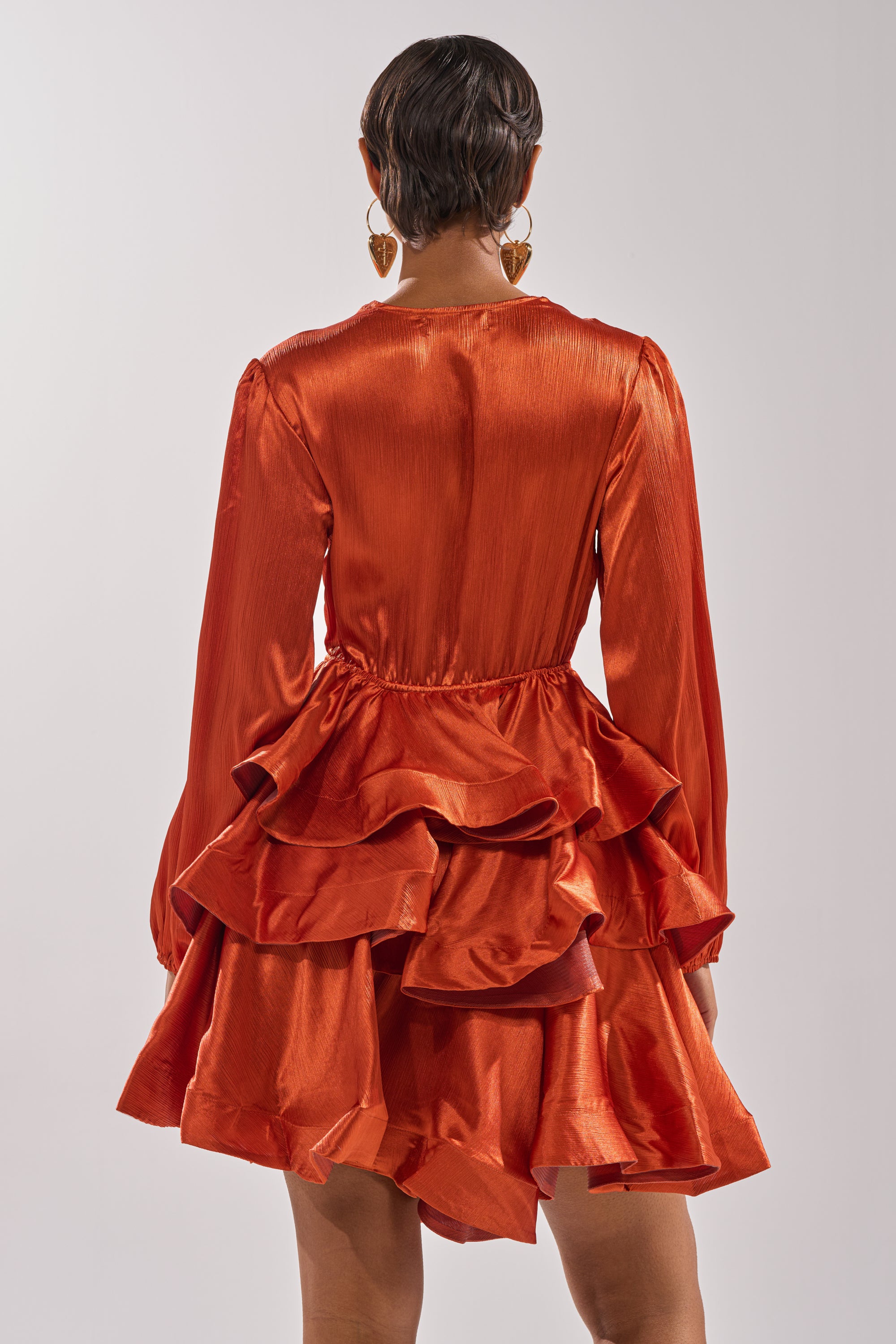 A person with short hair and gold earrings is seen from the back in the ARIANA SATIN RUFFLED MINI DRESS, featuring layered burnt orange ruffles, against a plain light background—ideal as chic evening wear.