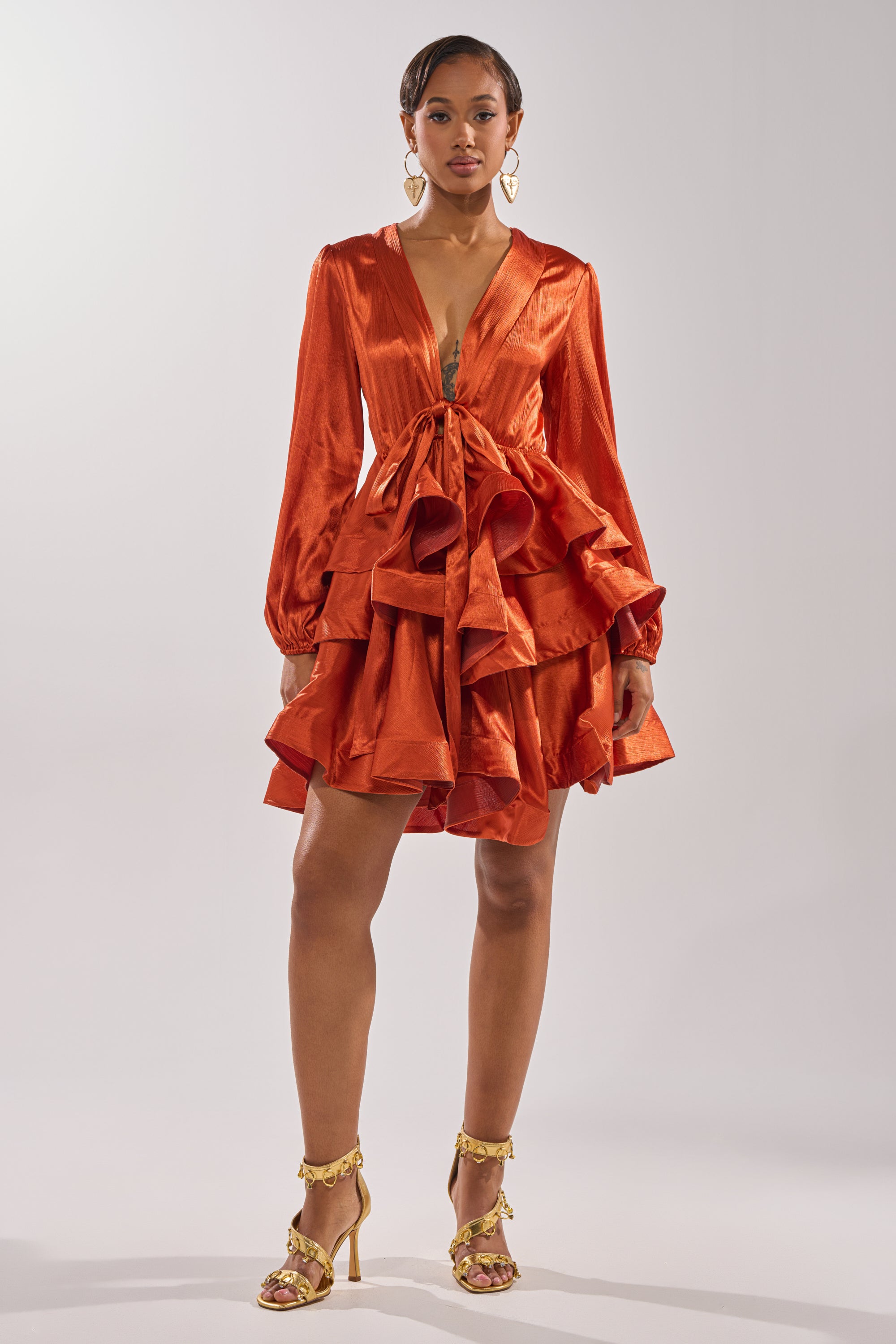 A woman models the ARIANA SATIN RUFFLED MINI DRESS in shiny burnt orange, featuring a deep V-neck and ruffled skirt, styled with gold heels and statement earrings—an elegant evening look against a plain background.