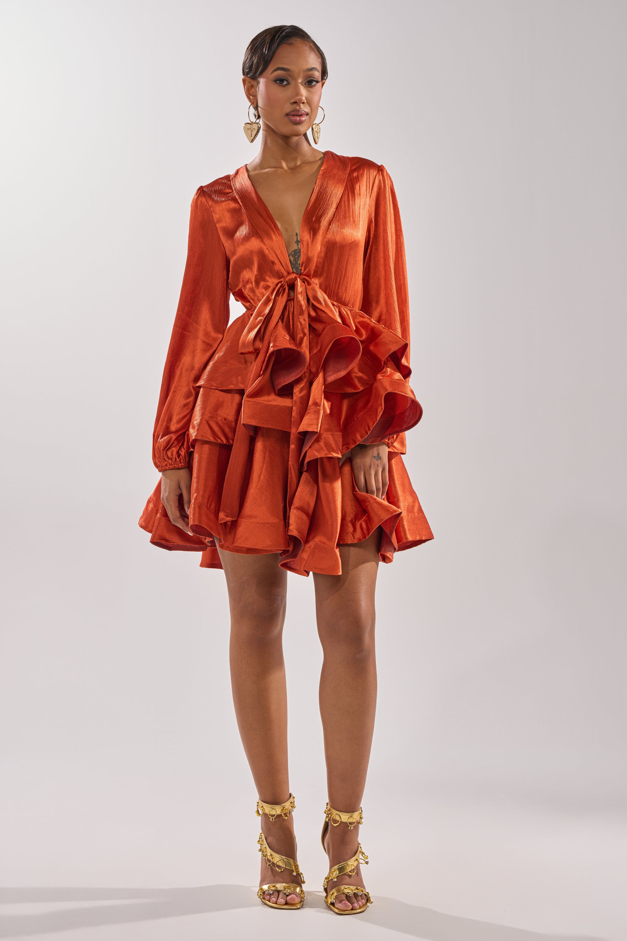 A woman wears the ARIANA SATIN RUFFLED MINI DRESS in shiny burnt orange, featuring long sleeves and a deep V-neckline, styled with gold heels and hoop earrings against a plain light background for a chic special occasion look.