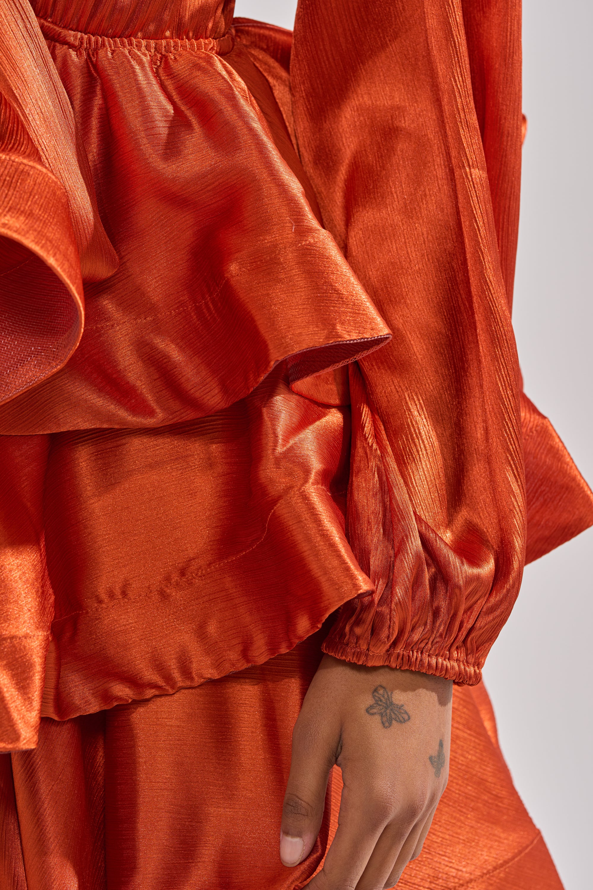 Close-up of a person wearing the ARIANA SATIN RUFFLED MINI DRESS in shiny burnt orange, featuring textured layers and long sleeves; their hand with a small butterfly tattoo rests by their side—ideal for special occasions.
