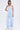 A woman models the SUCKER FOR YOU SEERSUCKER JUMPSUIT, featuring light blue and white duotone stripes, a wide leg silhouette, and cargo pockets, styled with white sneakers and silver necklaces against a plain white backdrop.