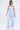 A woman stands confidently in the SUCKER FOR YOU SEERSUCKER JUMPSUIT—light blue, duotone striped, off-the-shoulder with wide legs and cargo pockets—paired with white sneakers and layered silver necklaces against a plain background.