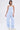 A woman poses against a plain background in the SUCKER FOR YOU SEERSUCKER JUMPSUIT, featuring a strapless neckline, light blue seersucker fabric, cargo pockets, and wide legs. She pairs it with white sneakers and layered silver necklaces.