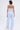 A woman with short dark hair faces away, wearing the SUCKER FOR YOU SEERSUCKER JUMPSUIT in light blue with a strapless neckline and wide legs, paired with white sneakers and a thin halter necklace against a white background.