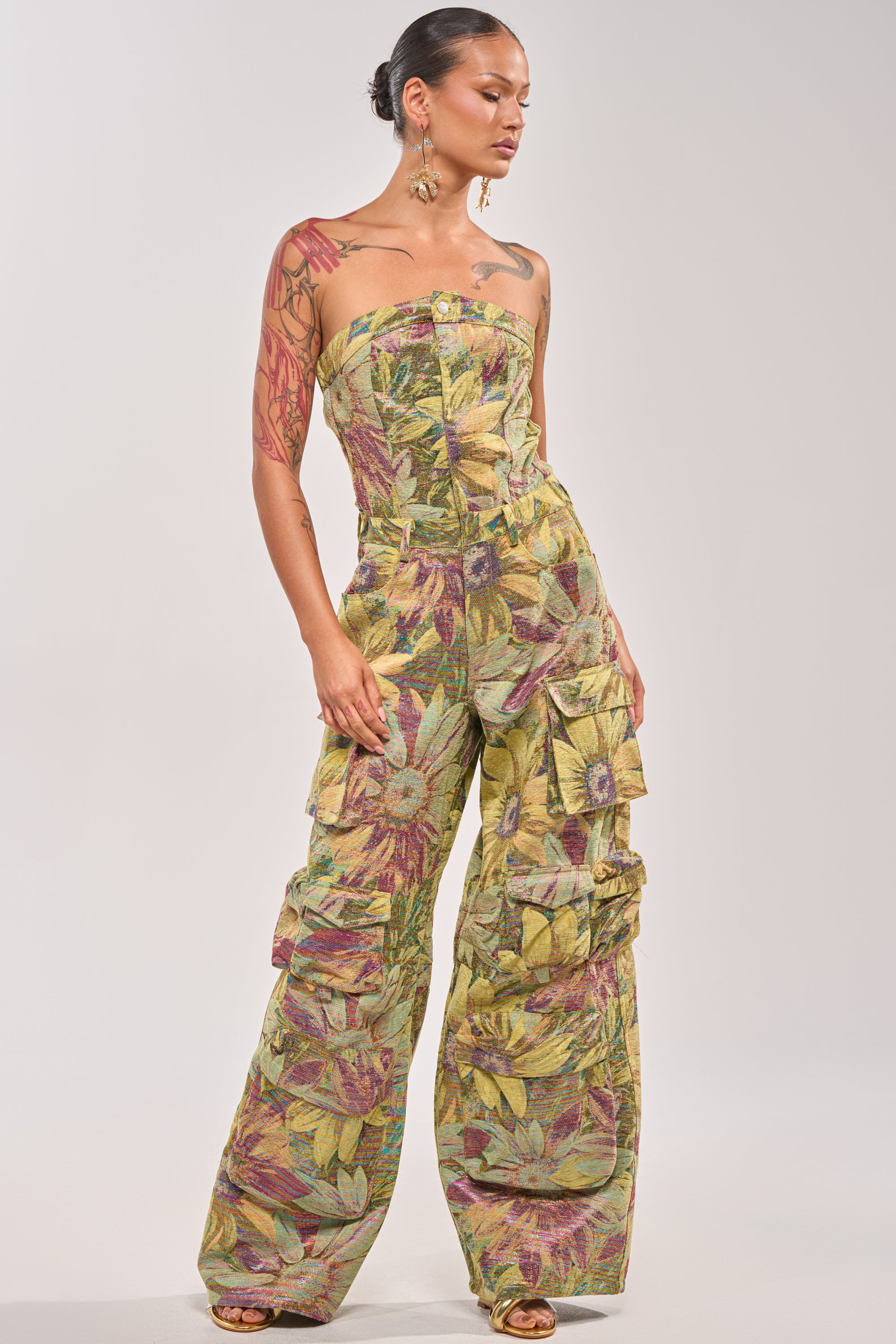 A person stands confidently against a plain background, wearing the SMELL THE FLOWERS BROCADE STRAPLESS JUMPSUIT featuring a colorful floral pattern and multiple cargo pockets. 0