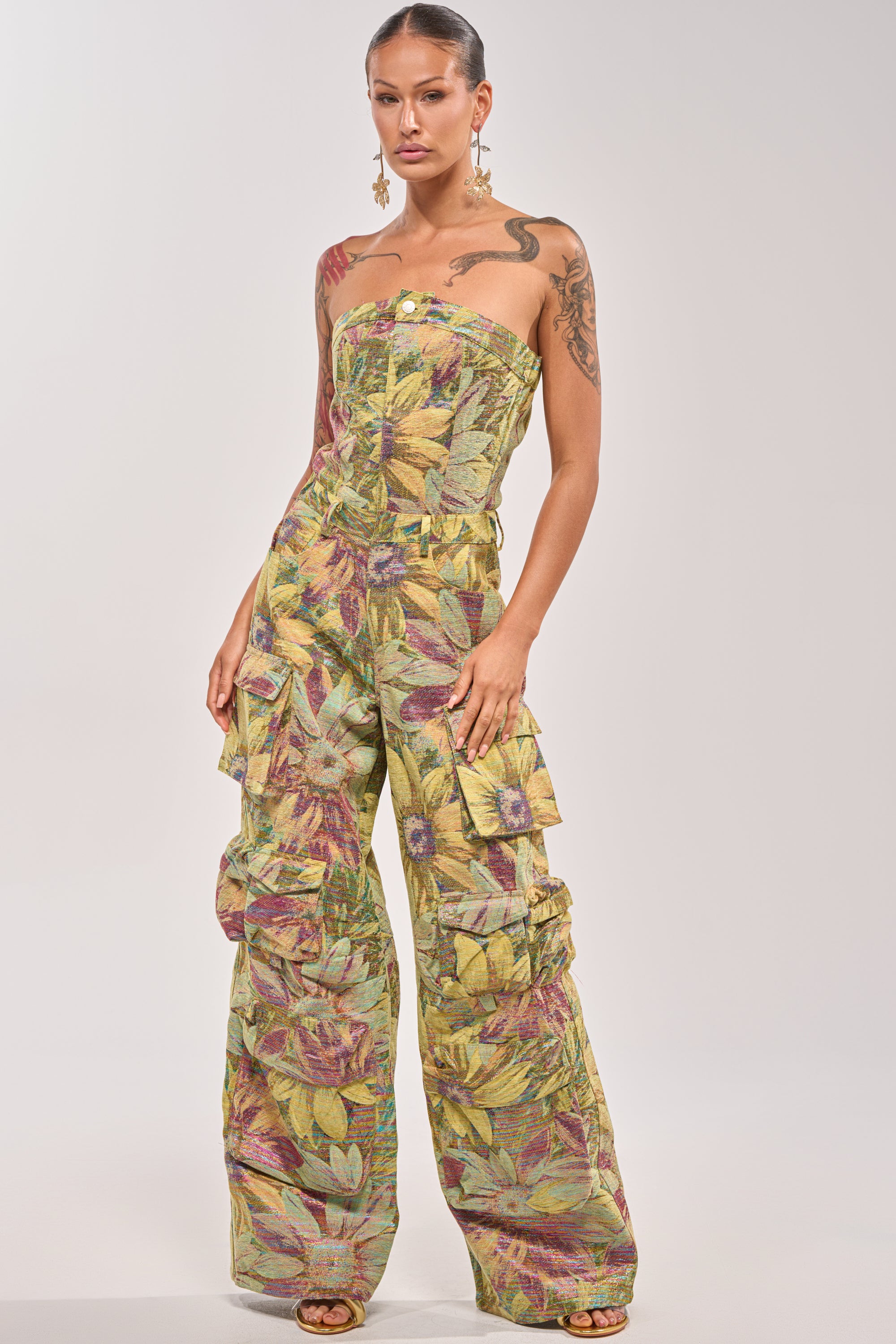A woman stands confidently in the SMELL THE FLOWERS BROCADE STRAPLESS JUMPSUIT, featuring a multicolored leafy floral print. She accessorizes with gold earrings, shows off her tattoos, and has her hair pulled back against a plain light background. 1
