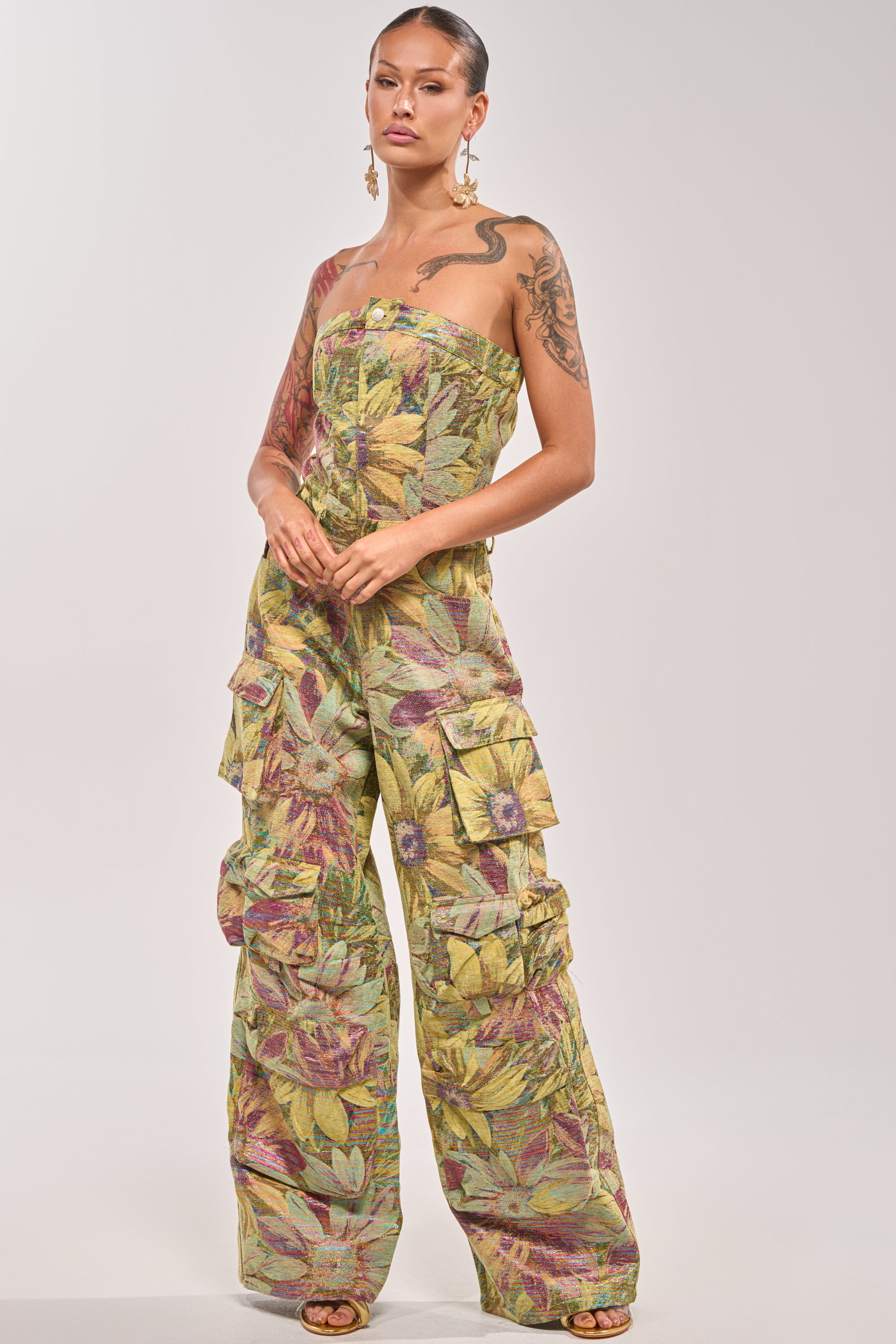 A woman with tattoos and slicked-back hair wears the SMELL THE FLOWERS BROCADE STRAPLESS JUMPSUIT, featuring a colorful leaf pattern, multiple cargo pockets, and statement earrings. 2