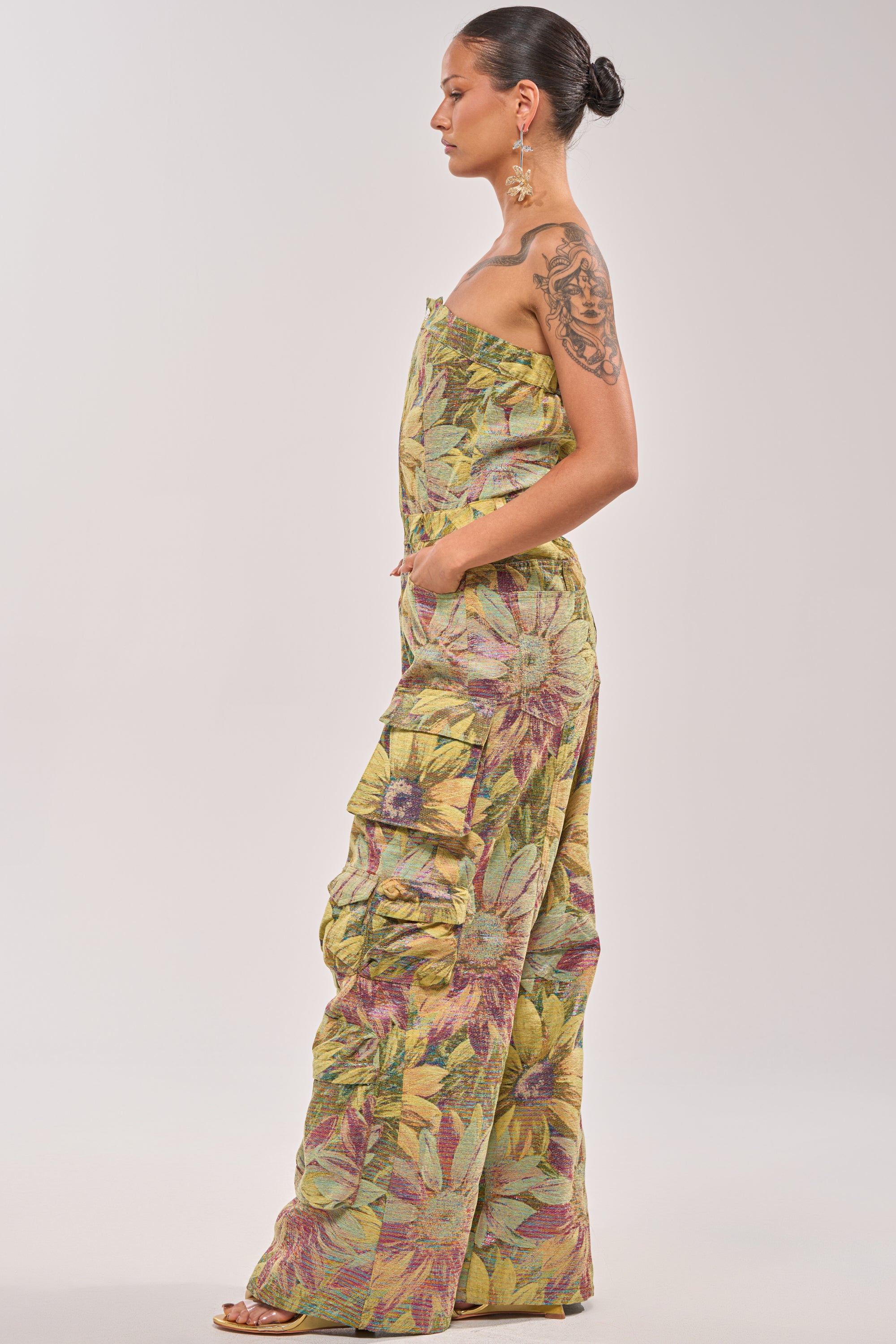 A woman in profile wears the SMELL THE FLOWERS BROCADE STRAPLESS JUMPSUIT featuring a yellow, green, and purple floral pattern. Her hair is in a bun, with large earrings and a visible tattoo on her upper arm. 3