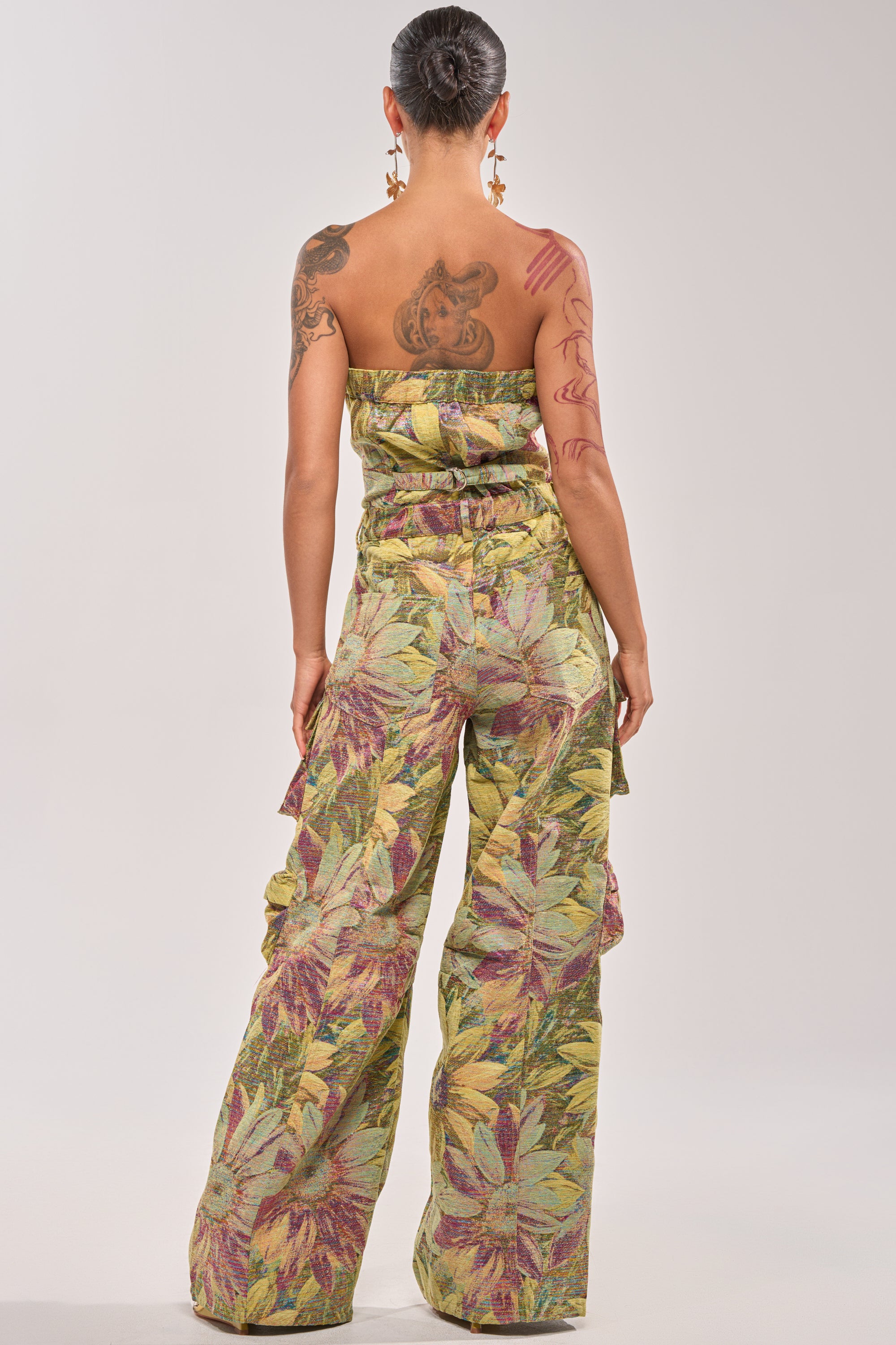 A tattooed person with star earrings is seen from behind, wearing the SMELL THE FLOWERS BROCADE STRAPLESS JUMPSUIT with green, yellow, and maroon leaf patterns against a plain background. 4
