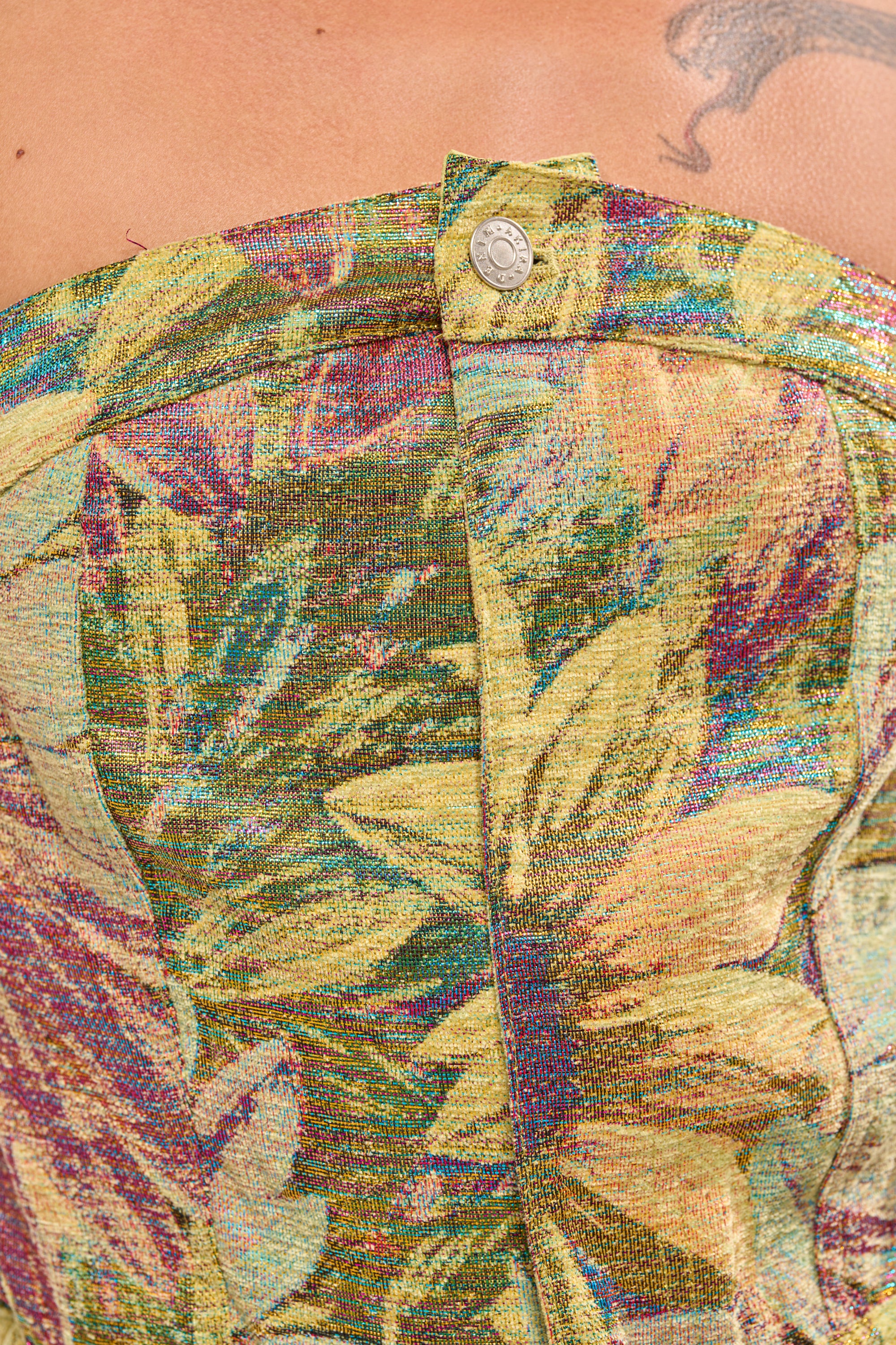 Close-up of the SMELL THE FLOWERS BROCADE STRAPLESS JUMPSUIT, featuring metallic floral-patterned fabric in yellow, green, and purple with a silver top button. A hint of tattooed skin is visible above the jumpsuit. 5