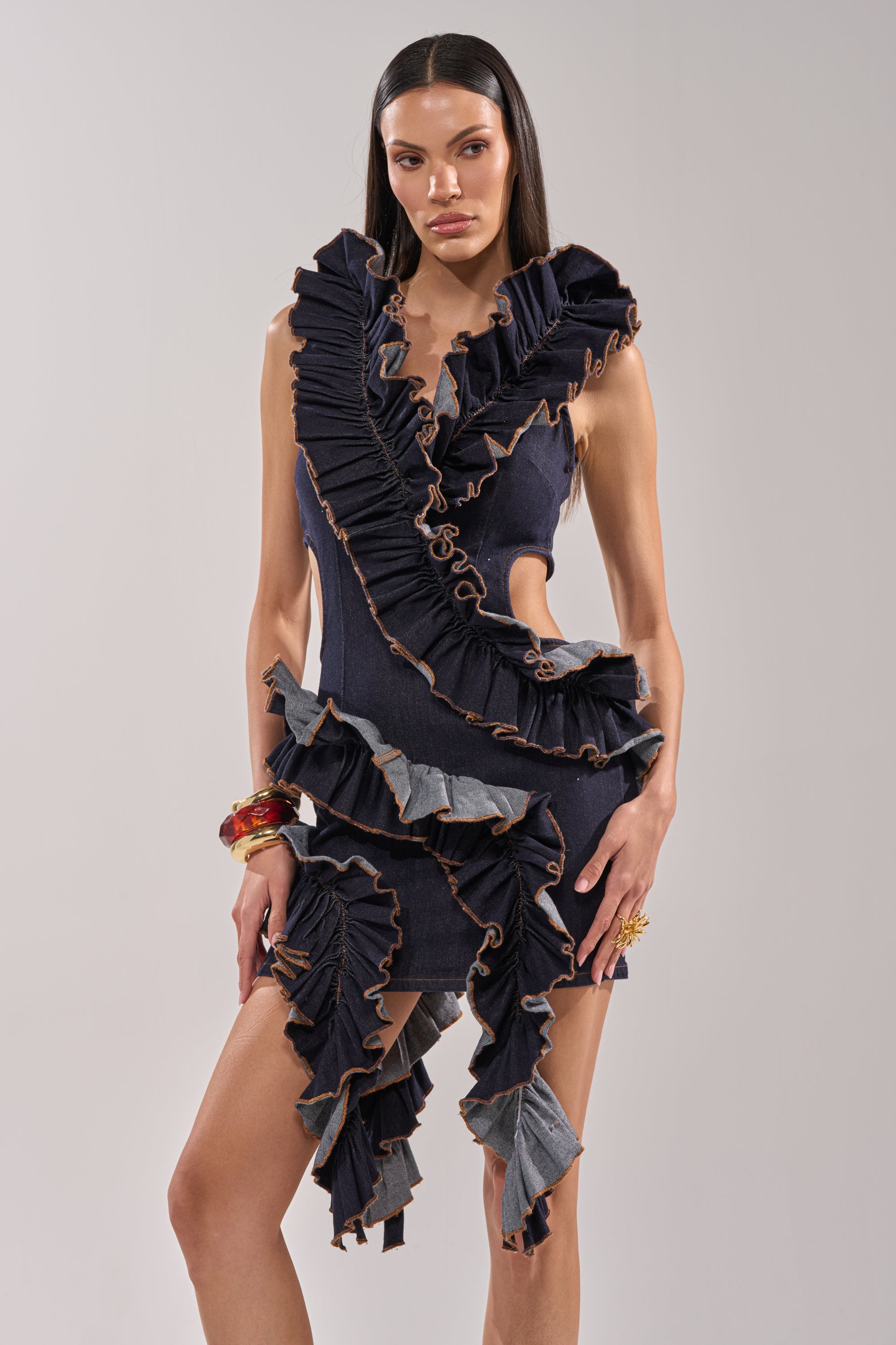 A woman wears the SAY IT AGAIN RUFFLED DENIM MINI DRESS IN DARK BLUE DENIM, featuring ruffle details, tan stitching, and dramatic cutouts. She pairs it with statement jewelry for a chic, modern look. 2