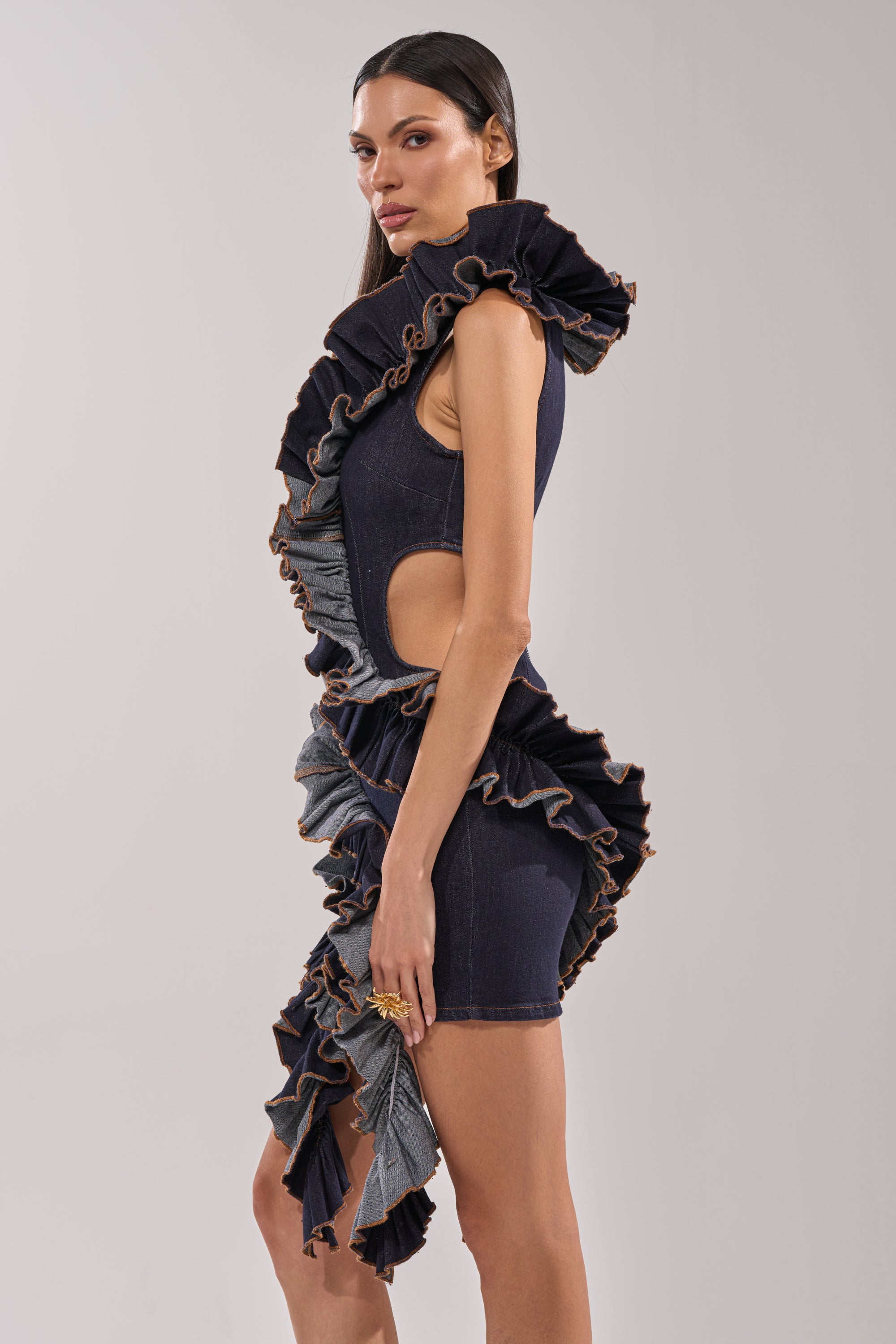 A woman stands in profile against a light gray background, wearing the SAY IT AGAIN RUFFLED DENIM MINI DRESS IN DARK BLUE DENIM, featuring cutouts and dramatic ruffle detailing at the edges. 3