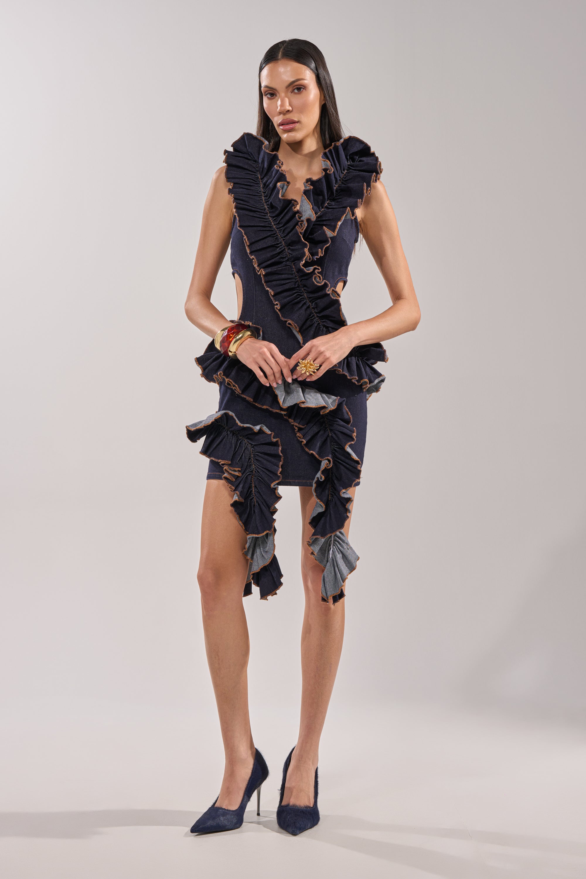 A woman stands in a studio wearing the SAY IT AGAIN RUFFLED DENIM MINI DRESS IN DARK BLUE DENIM, a sleeveless bodycon mini with asymmetrical ruffled layers and orange trim, paired with matching high heels and minimal accessories. 5