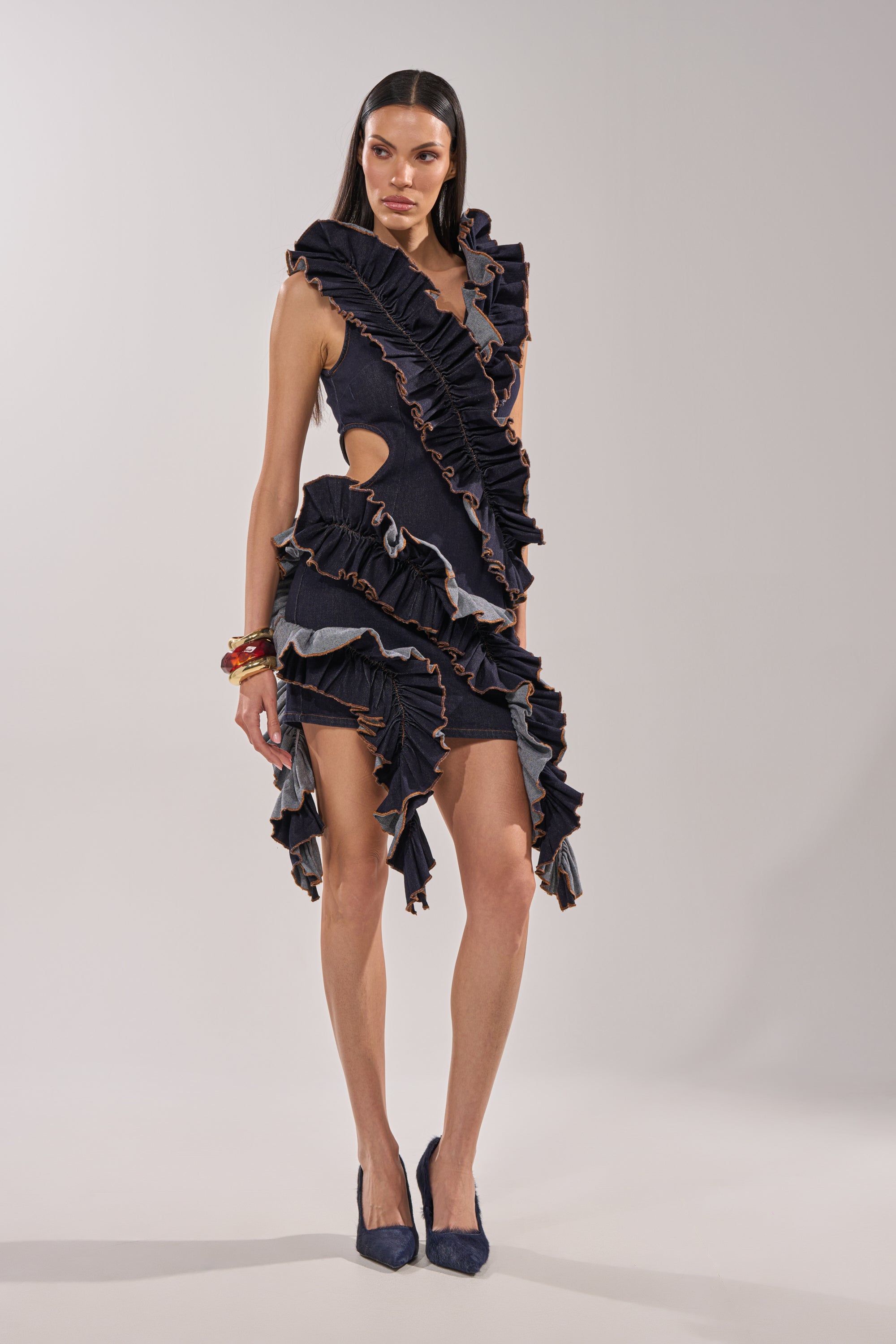 A woman poses in a studio wearing the SAY IT AGAIN RUFFLED DENIM MINI DRESS IN DARK BLUE DENIM, featuring sleeveless cutouts and bold ruffle details. She styles it with straight dark hair, stacked bracelets, and pointed dark heels. 6