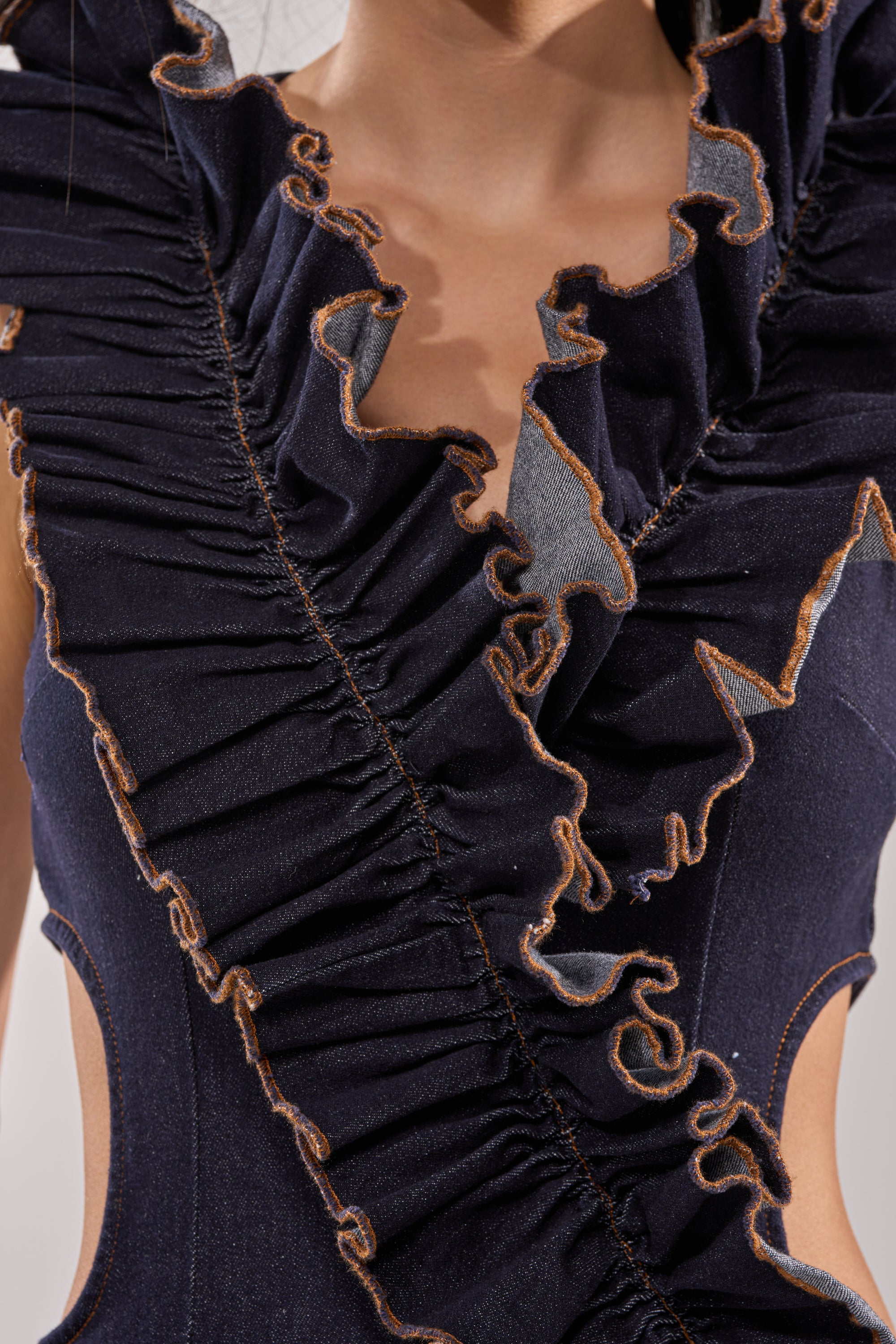 Close-up of a person wearing the SAY IT AGAIN RUFFLED DENIM MINI DRESS IN DARK BLUE DENIM, featuring ruffle accents, exposed brown-edged seams, cut-out details, and intricate textured fabric near the chest and shoulders. 7