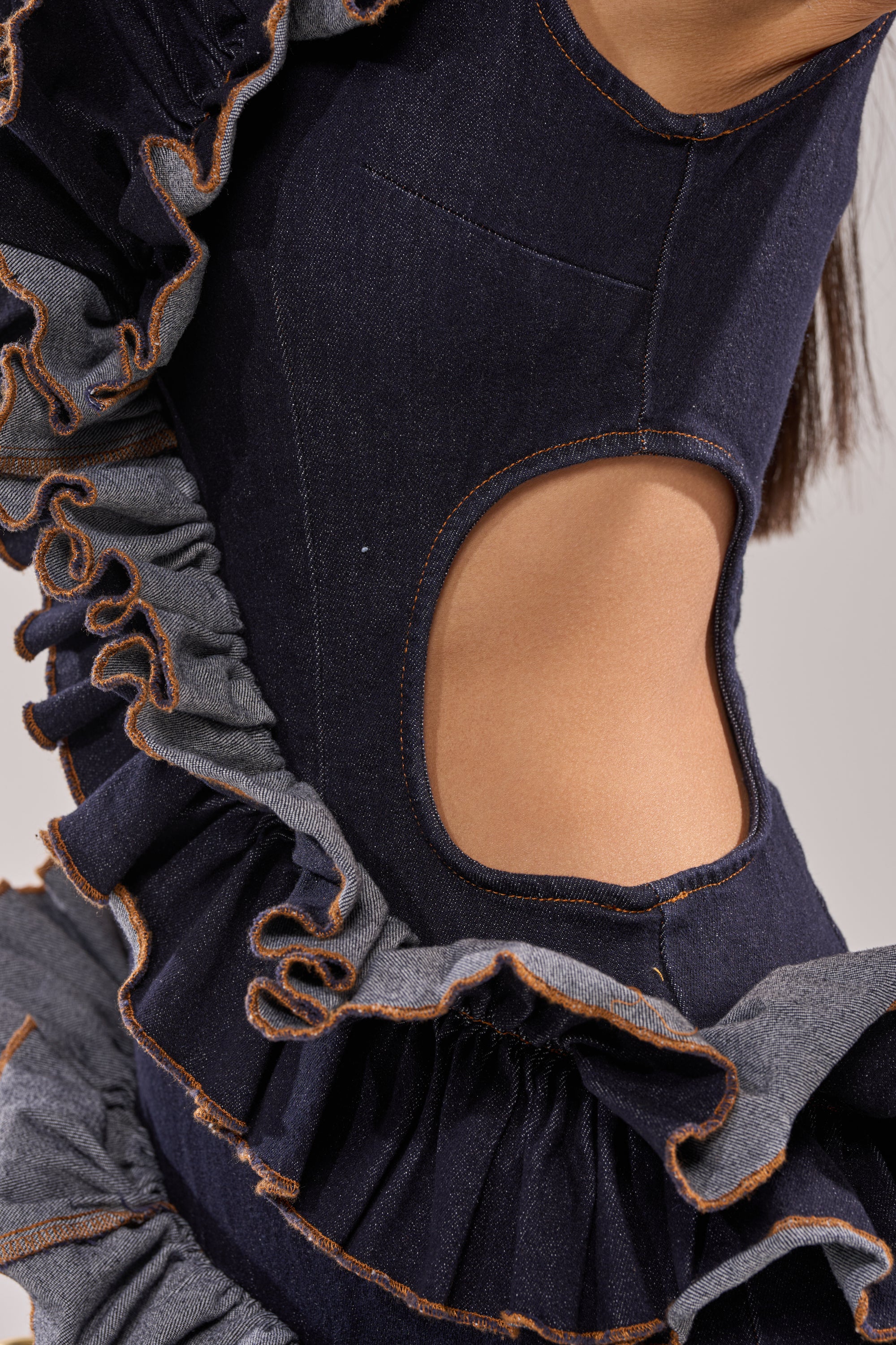 A close-up of a person wearing the SAY IT AGAIN RUFFLED DENIM MINI DRESS IN DARK BLUE DENIM, featuring a large circular waist cutout and contrasting gray ruffle detailing with brown stitching. 8