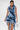 A woman models the SAY IT AGAIN RUFFLED DENIM MINI DRESS, featuring dramatic layered frills, side cutouts, and an asymmetric hemline. She holds a small silver handbag against a plain white background with her sleek dark hair pulled back.