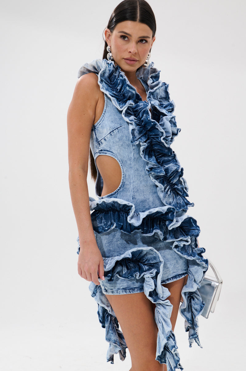 A woman models the SAY IT AGAIN RUFFLED DENIM MINI DRESS with bold ruffled edges and side cutouts in layered blue denim, pairing it with statement earrings and a silver bag. Her long dark hair flows against a white backdrop.
