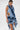 A woman models the SAY IT AGAIN RUFFLED DENIM MINI DRESS with bold ruffled edges and side cutouts in layered blue denim, pairing it with statement earrings and a silver bag. Her long dark hair flows against a white backdrop.