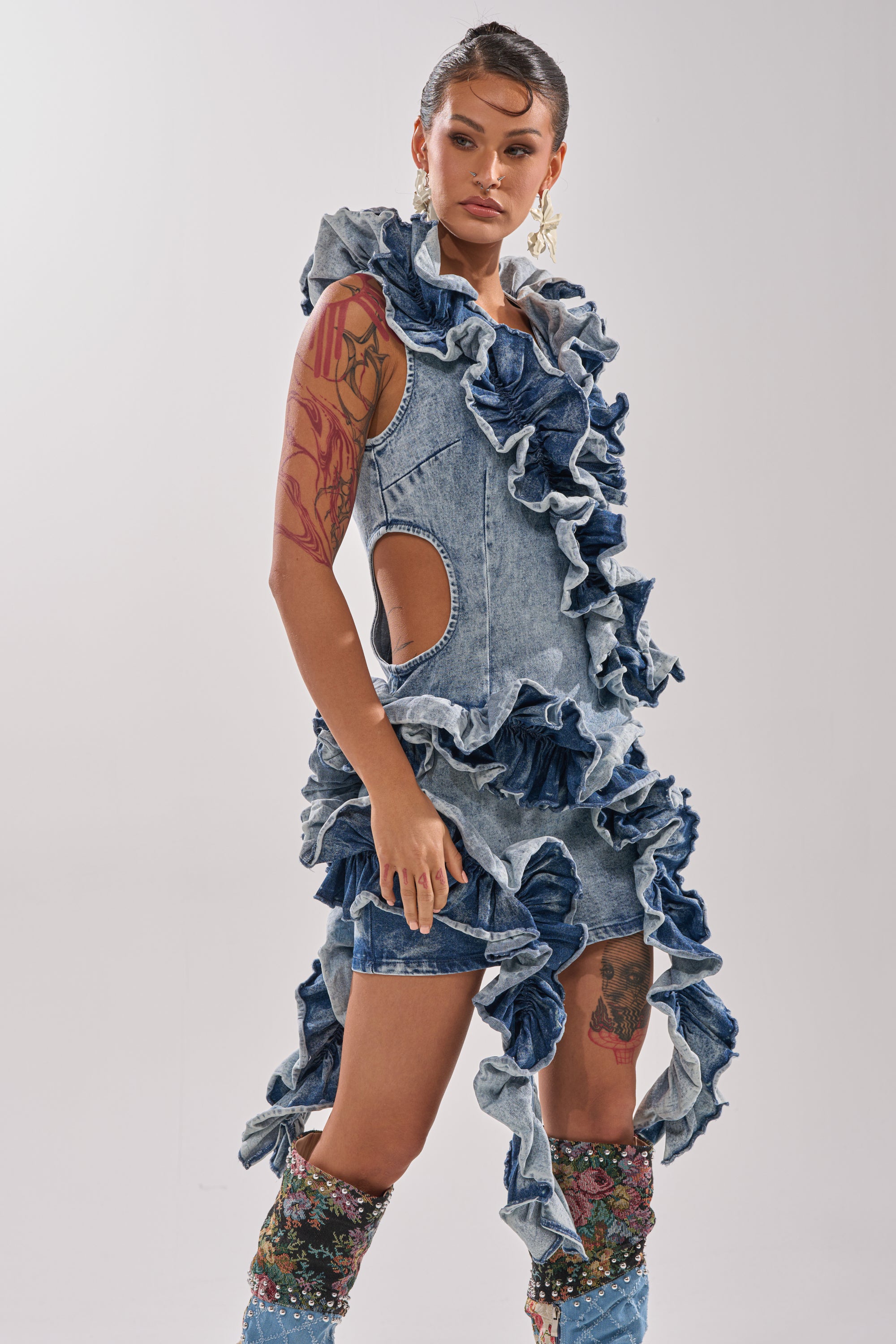 Wearing the SAY IT AGAIN RUFFLED DENIM MINI DRESS with side cutouts and matching ruffles, a person with visible arm tattoos and colorful boots stands confidently against a plain, light background. 4