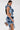 A woman with long dark hair models the SAY IT AGAIN RUFFLED DENIM MINI DRESS, featuring blue and white accents and side cutouts. She holds a silver clutch and poses against a plain white background.